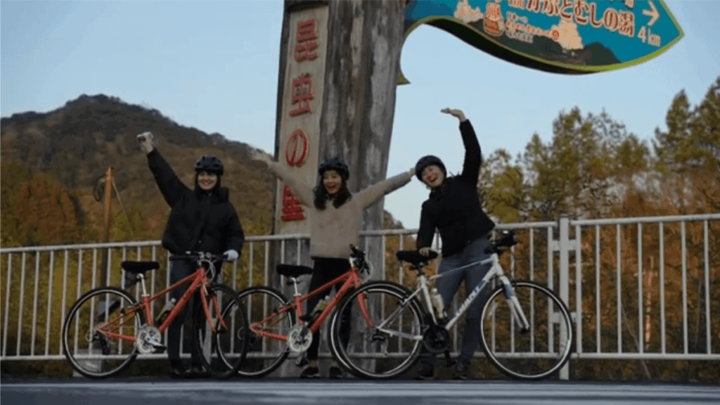 昆虫の館の縦看板前で、自転車を降りて並ぶ3人。背景に山々が広がる自然の風景